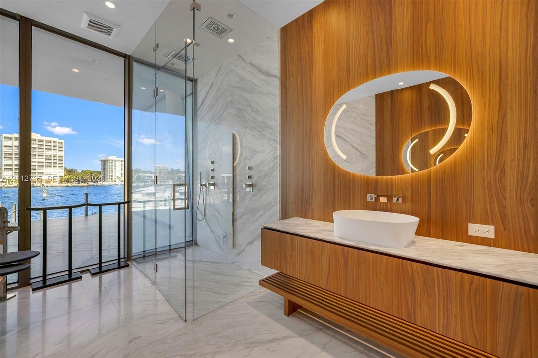 Floating wood-finish vanity with a stone countertop and vessel sink, illuminated oval mirror, walk-in glass-enclosed shower, stone-surround shower walls, and full-height floor-to-ceiling windows