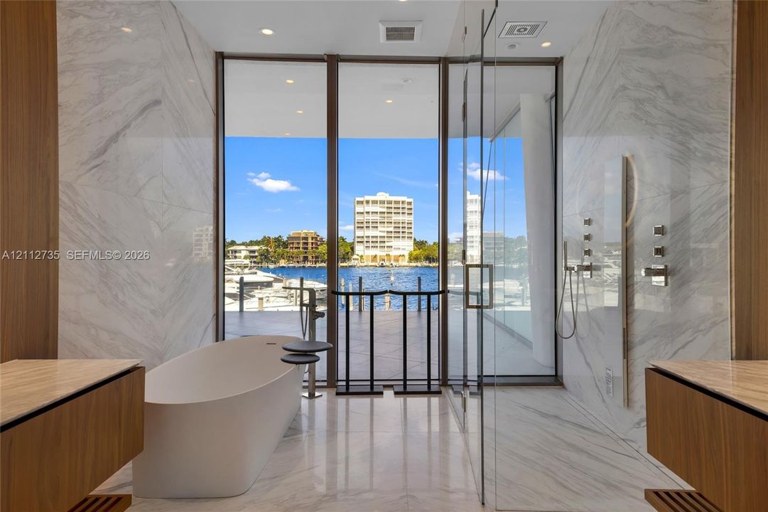 Floor-to-ceiling glass windows, stone-finish wall cladding, freestanding soaking tub, glass-enclosed shower, and dual wood-finish floating vanities