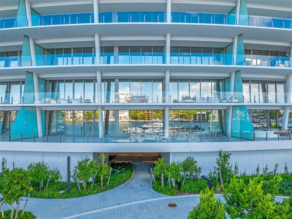 Contemporary multi-story facade featuring floor-to-ceiling glass windows, wraparound balconies with glass railings, structural white pillars, and landscaped ground-level pavers