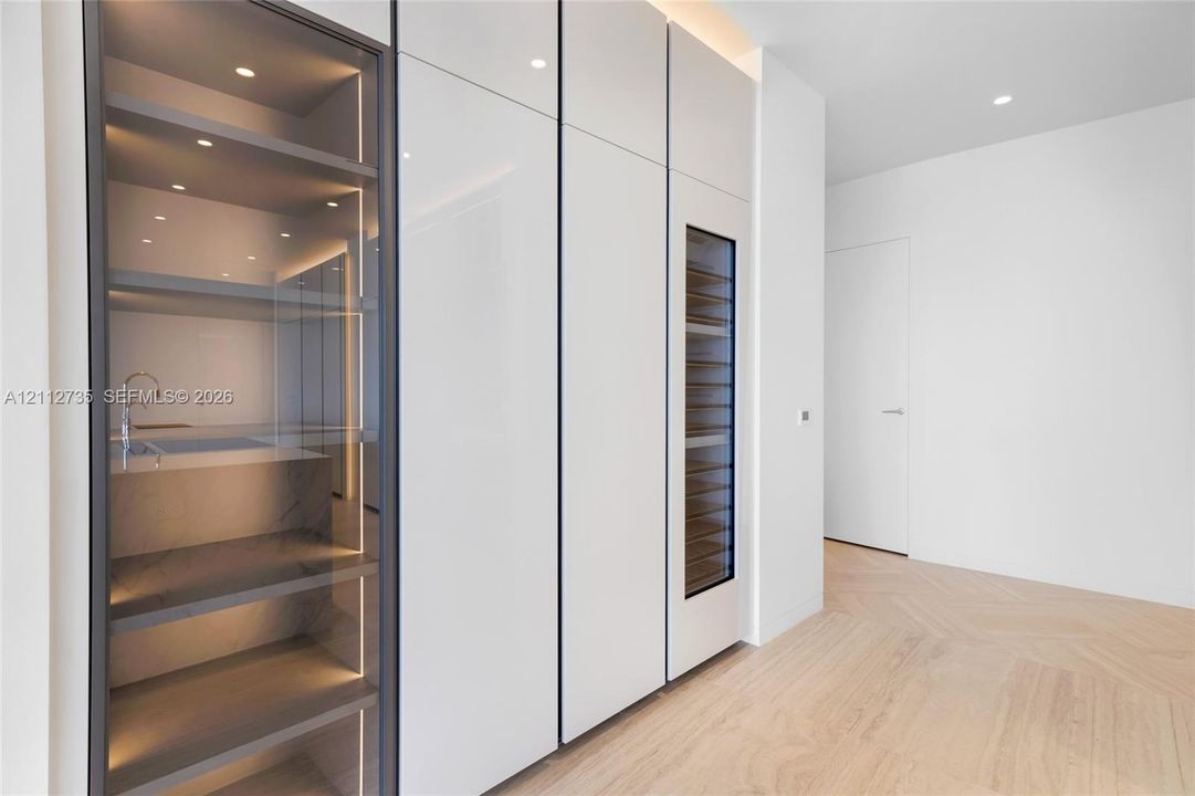 Custom millwork featuring floor-to-ceiling high-gloss cabinetry, integrated wine refrigeration, glass-front display shelving with LED illumination, and chevron wood-finish flooring