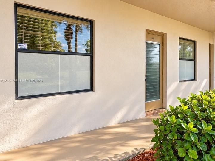 Exterior facade featuring a smooth stucco finish, single-pane windows with dark frames, and a glass-paneled entry door with horizontal louvers
