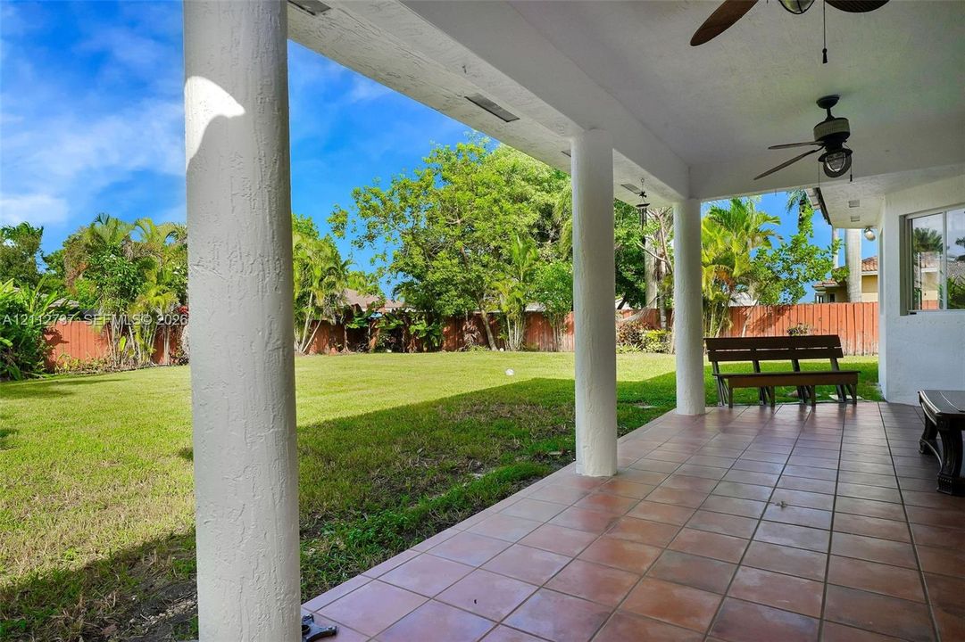 Covered patio featuring ceramic tile flooring, structural pillars, and ceiling-mounted fans overlooking a fenced backyard with mature trees