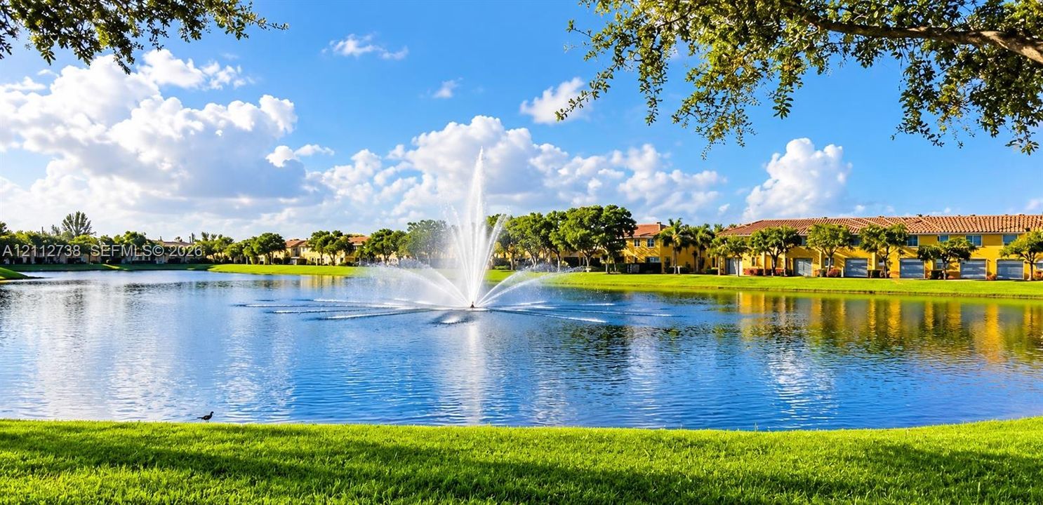 Large community lake featuring a central aerating fountain, manicured lawn perimeter, and mature trees