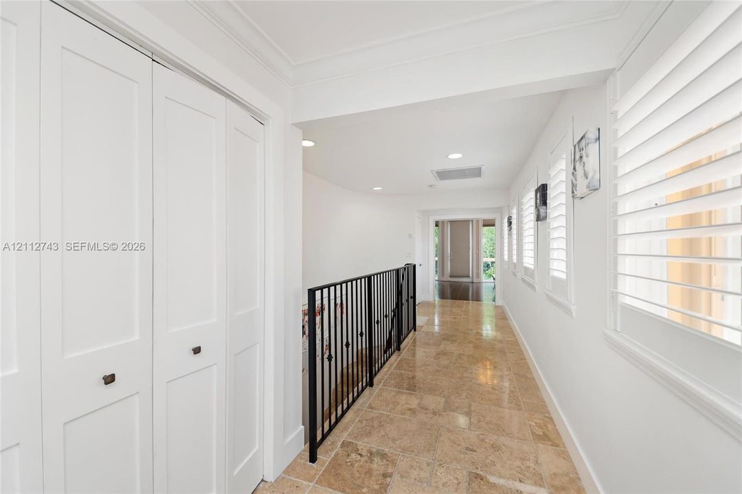 Hallway featuring stone tile flooring, white bi-fold closet doors, recessed lighting, plantation shutters, and a wrought-iron railing