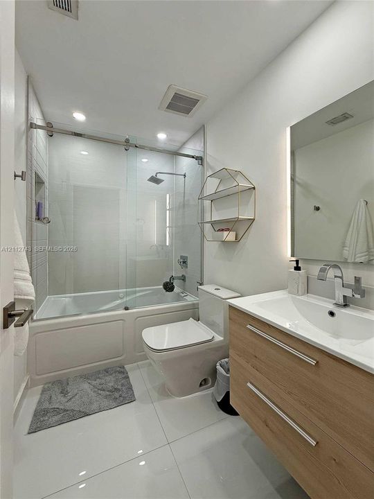 Bathroom featuring a wood-finish floating vanity with a white integrated sink, glass-enclosed shower and tub combination with a rain-style showerhead, recessed wall niche, and large-format tile flooring