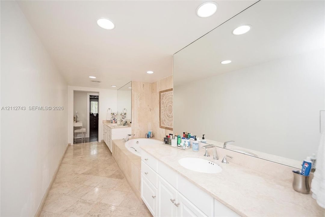 Bathroom featuring stone-finish flooring, a jetted soaking tub with a tiled surround, a lengthy stone-surface vanity with dual integrated sinks, and expansive wall-to-wall mirrors