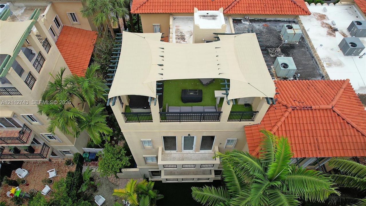Rooftop terrace featuring synthetic turf, metal railings, and a tensile shade canopy, complemented by multiple lower-level balconies and terracotta roof tiles