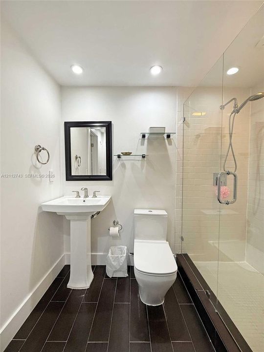 Pedestal sink with chrome widespread faucet, walk-in shower featuring a glass enclosure and wall-mounted hand shower, dark tile flooring, and integrated glass shelving