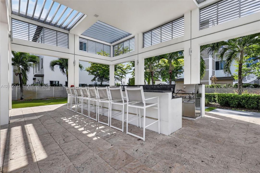 Covered outdoor kitchen featuring a stone-topped bar, stainless steel grill, horizontal slat ceiling panels, and natural stone paver flooring