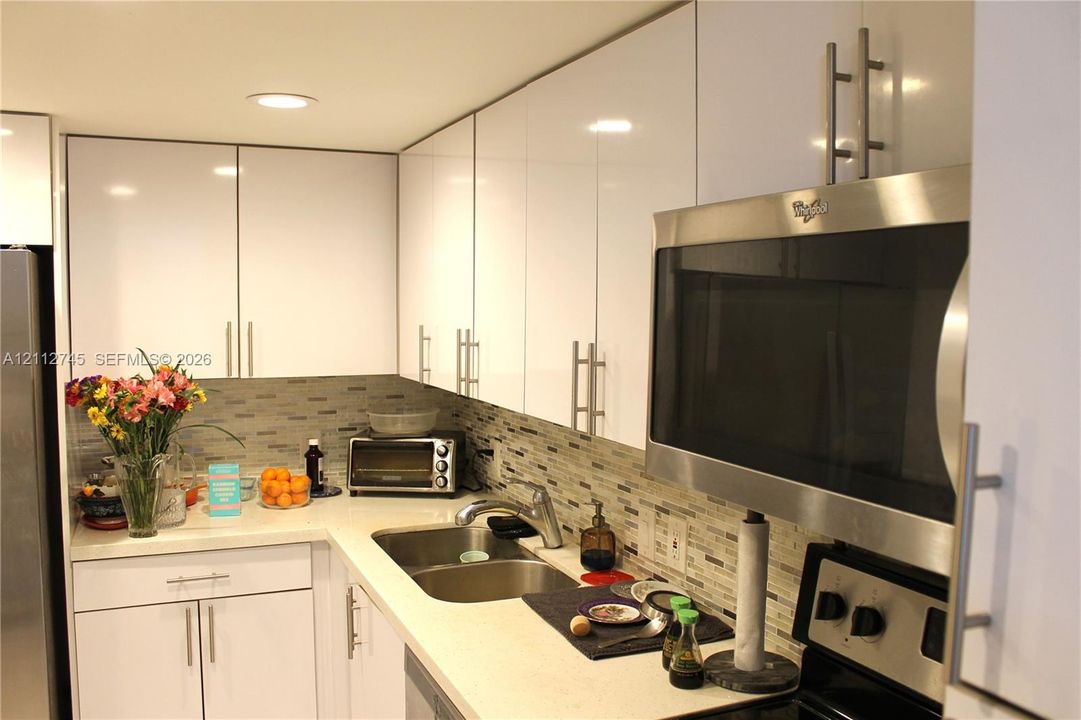 Glossy white cabinetry, solid-surface countertops, mosaic tile backsplash, recessed ceiling lighting, and stainless steel over-the-range microwave