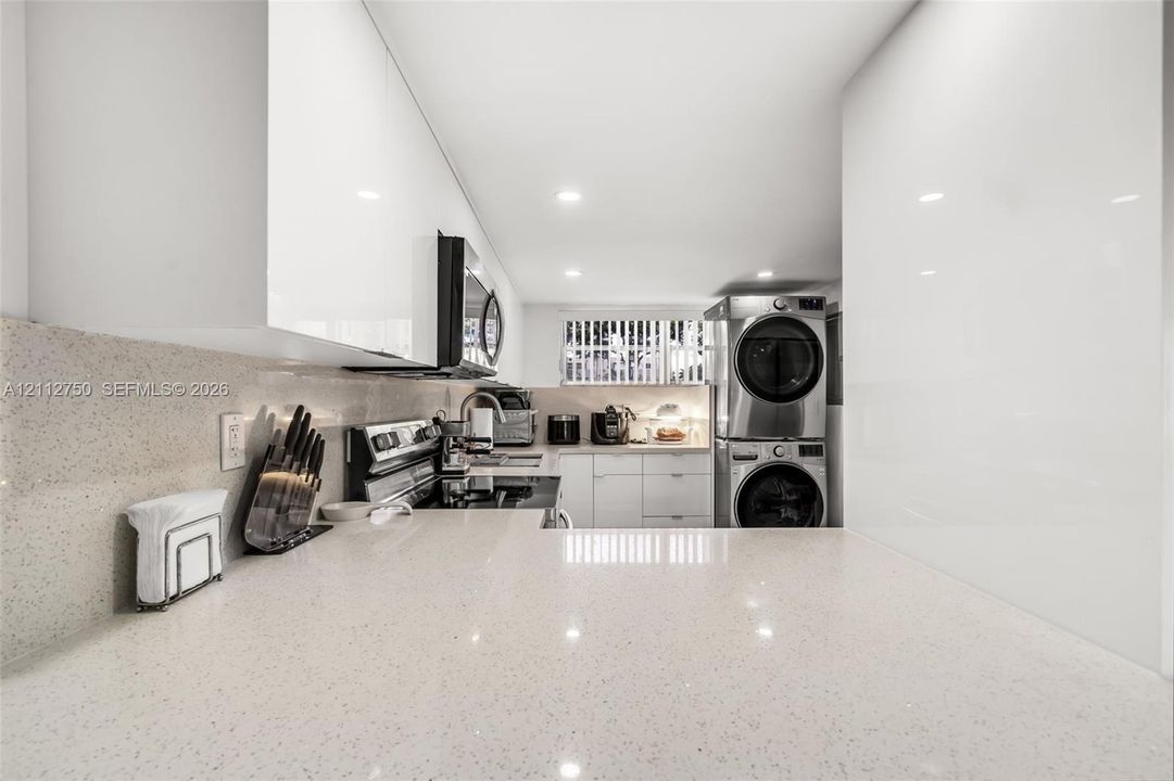 Kitchen area featuring gloss-finish cabinetry, stone-texture countertops, stacked stainless steel laundry appliances, and recessed ceiling lighting