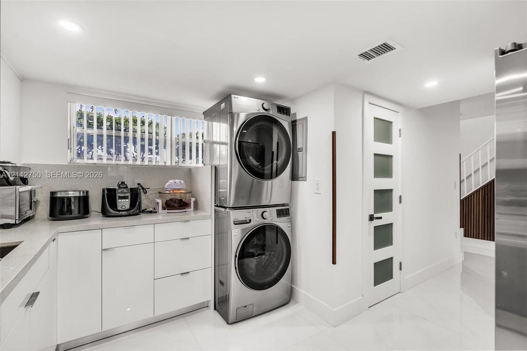 Laundry room featuring high-gloss white cabinetry, stone-finish countertops, stacked stainless steel appliances, and a frosted-glass panel door