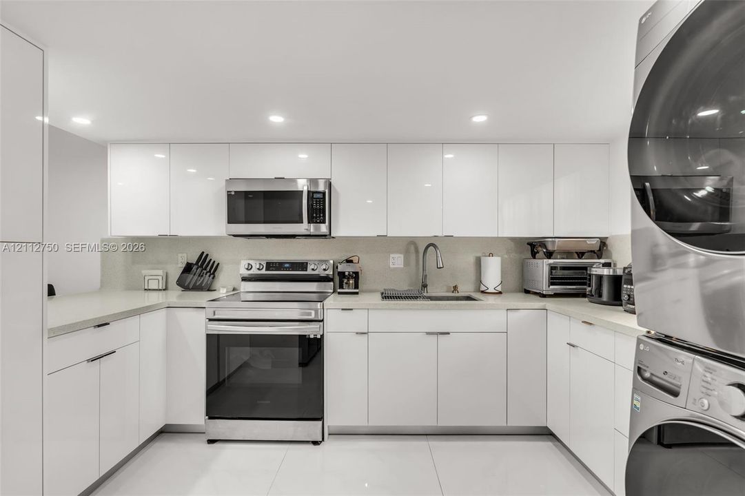 Modern kitchen featuring high-gloss white cabinetry, neutral stone-finish countertops, stainless steel appliances, a pull-down kitchen faucet, and large-format white tile flooring