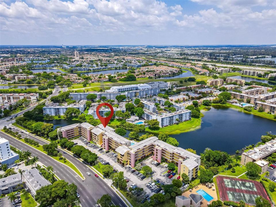 Aerial perspective of a multi-story residential building featuring on-site surface parking, a swimming pool, and a tennis court, situated on a lakefront lot