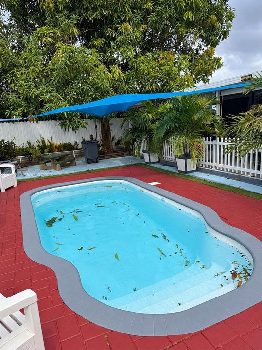 In-ground pool with molded steps, surrounded by red brick-patterned paving and a gray bullnose coping border, complemented by white picket fencing and a blue overhead tension shade sail