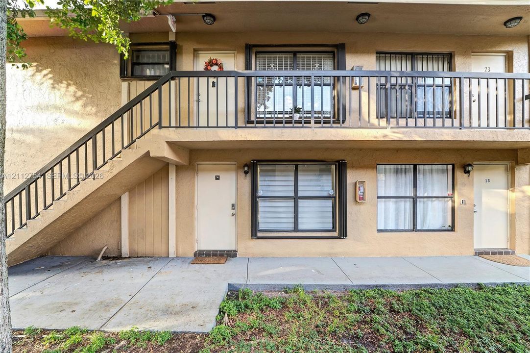 Exterior featuring a stucco facade, covered concrete walkway, second-level balcony with metal railing, and an external staircase