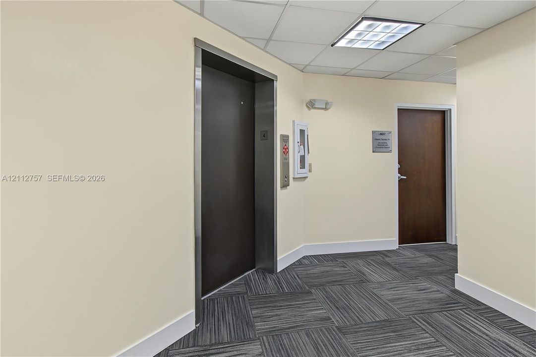 Elevator entry featuring a metal-finish door, industrial-style carpet tiles, drop ceiling with recessed lighting, and a solid-core wooden door