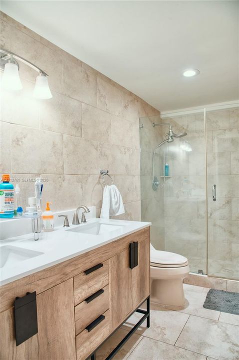 Double vanity featuring a white countertop with dual integrated sinks and a wood-finish cabinet base, complemented by a walk-in glass-enclosed shower with neutral tile surround