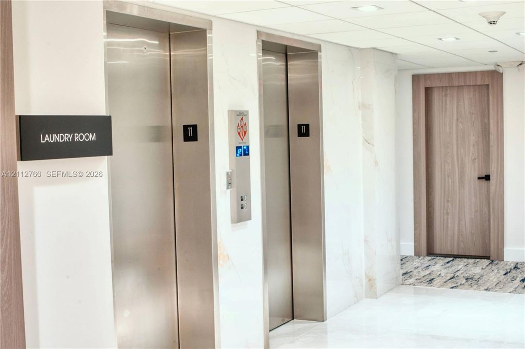 Elevator lobby featuring stainless steel doors, white stone-finish wall cladding, recessed ceiling lighting, and a wood-finish interior door