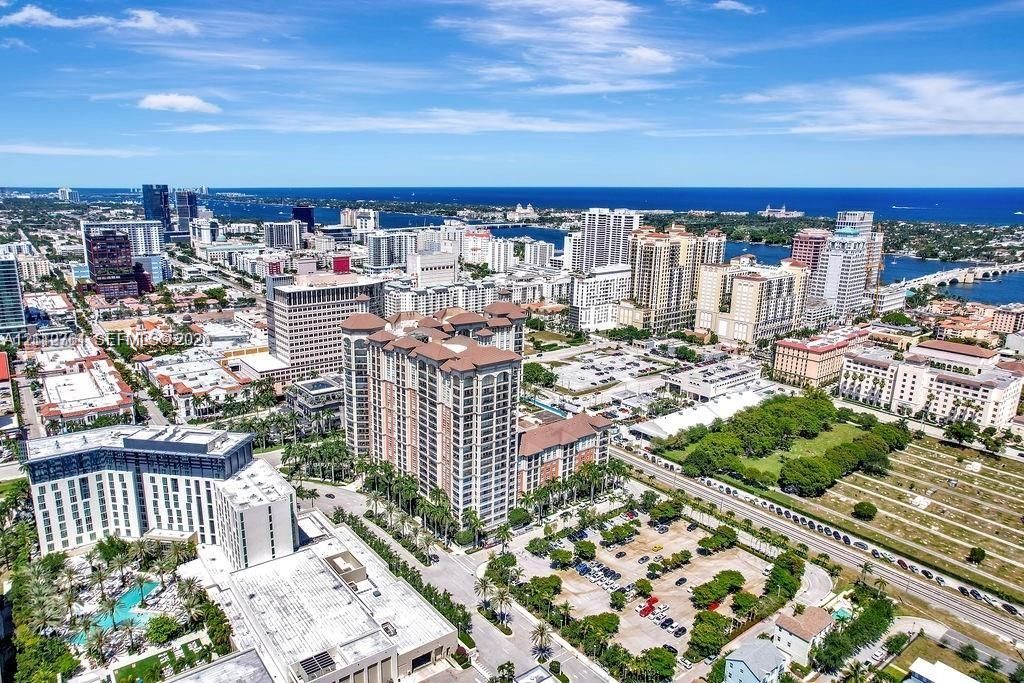 Aerial perspective featuring a high-rise residential structure, urban cityscape, dense foliage, paved parking areas, and a coastal waterway backdrop