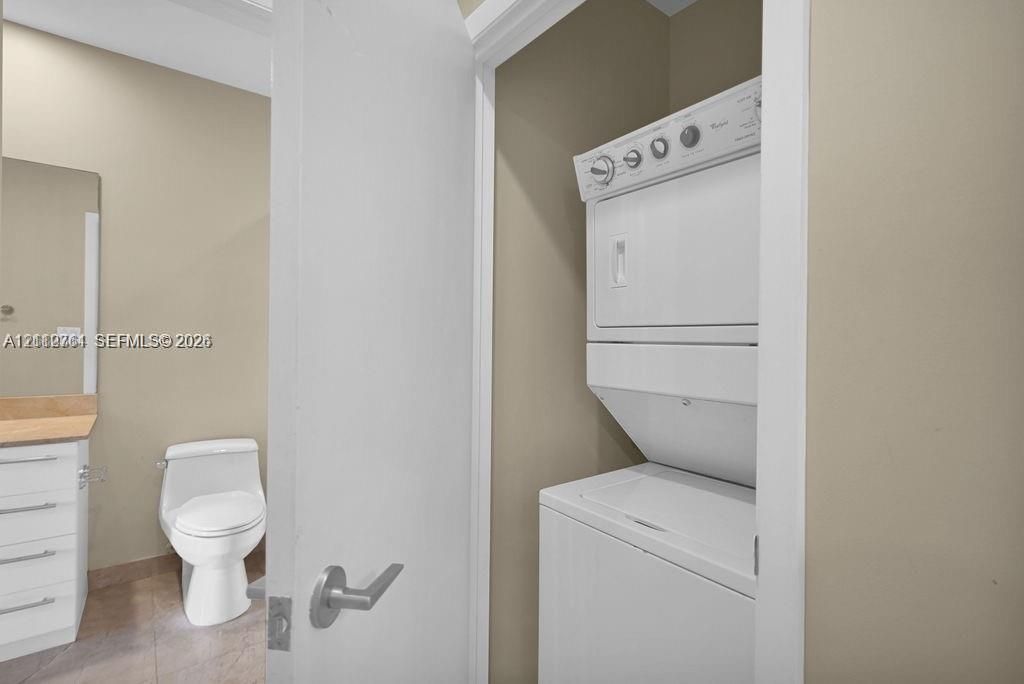 Laundry closet featuring a white stacked washer and dryer unit, neutral-toned walls, a white vanity with a neutral stone-finish countertop, and a white porcelain commode
