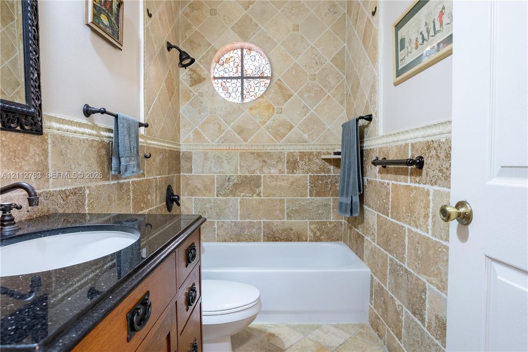 Bathroom featuring natural stone tile walls in a combination of brick-set and diagonal patterns, dark stone-finish vanity countertop, wood-finish cabinetry, built-in recessed wall niche, and circular accent window