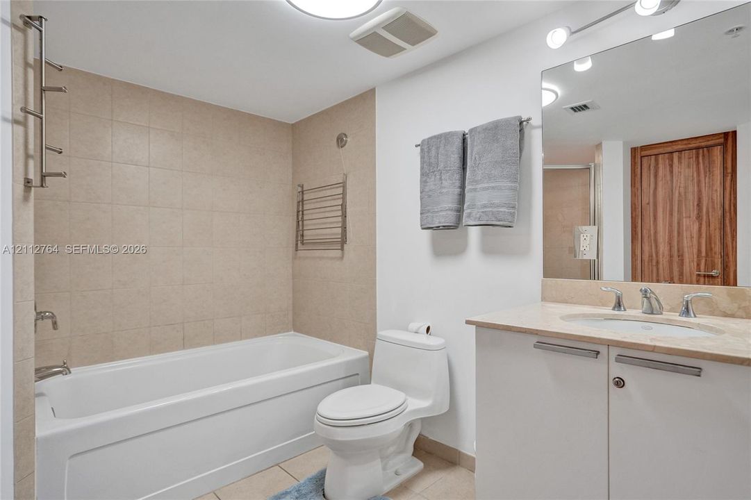 Bathroom featuring a deep soaking tub with neutral tile surround, vanity with stone-finish countertop, undermount sink, and full-width vanity mirror with integrated lighting