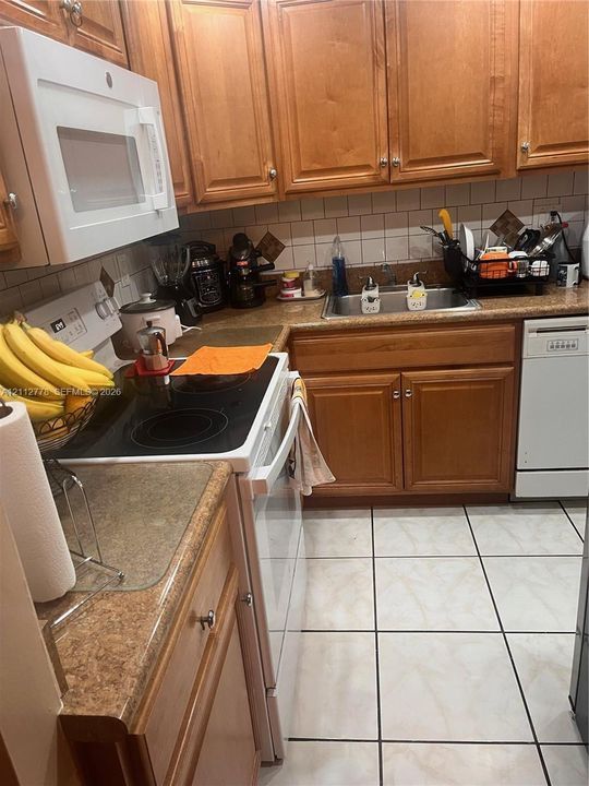 Kitchen featuring natural wood cabinetry, white tile backsplash, stone-finish countertops, ceramic tile flooring, and integrated white appliances