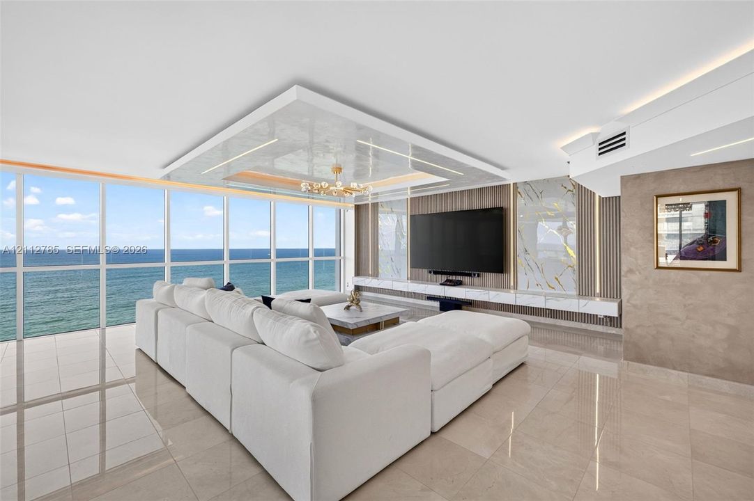 Floor-to-ceiling windows featuring ocean scenery, expansive polished tile flooring, tray ceiling with recessed lighting, and a built-in media wall with stone-finish paneling and under-cabinet illumination