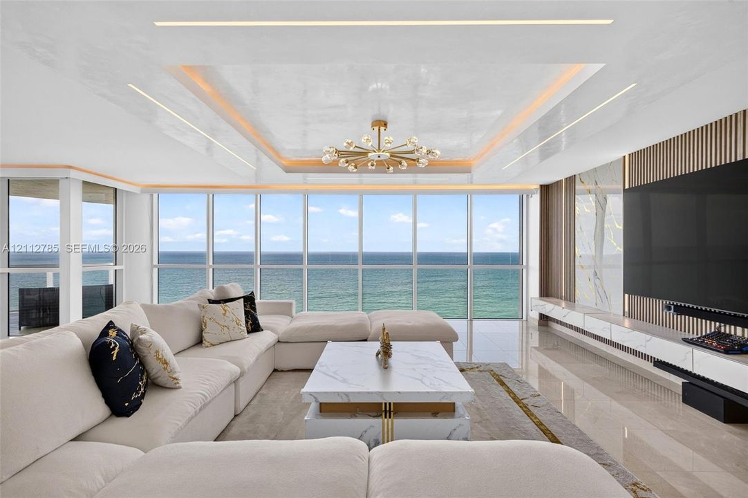 Floor-to-ceiling windows featuring ocean scenery, tray ceiling with recessed cove lighting, central sputnik-style chandelier, and custom built-in media wall with stone cladding and vertical wood slat detailing