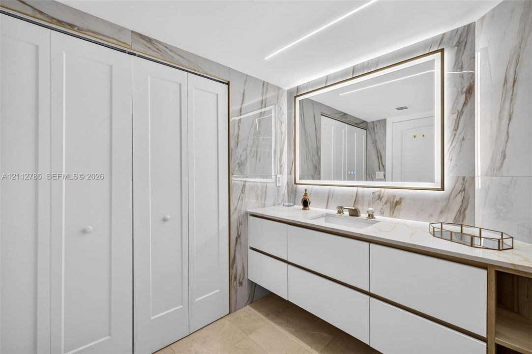 Floating vanity featuring flat-panel drawers, light-toned stone-finish countertop, integrated under-mount sink, LED-backlit mirror, and floor-to-ceiling porcelain tile wall cladding