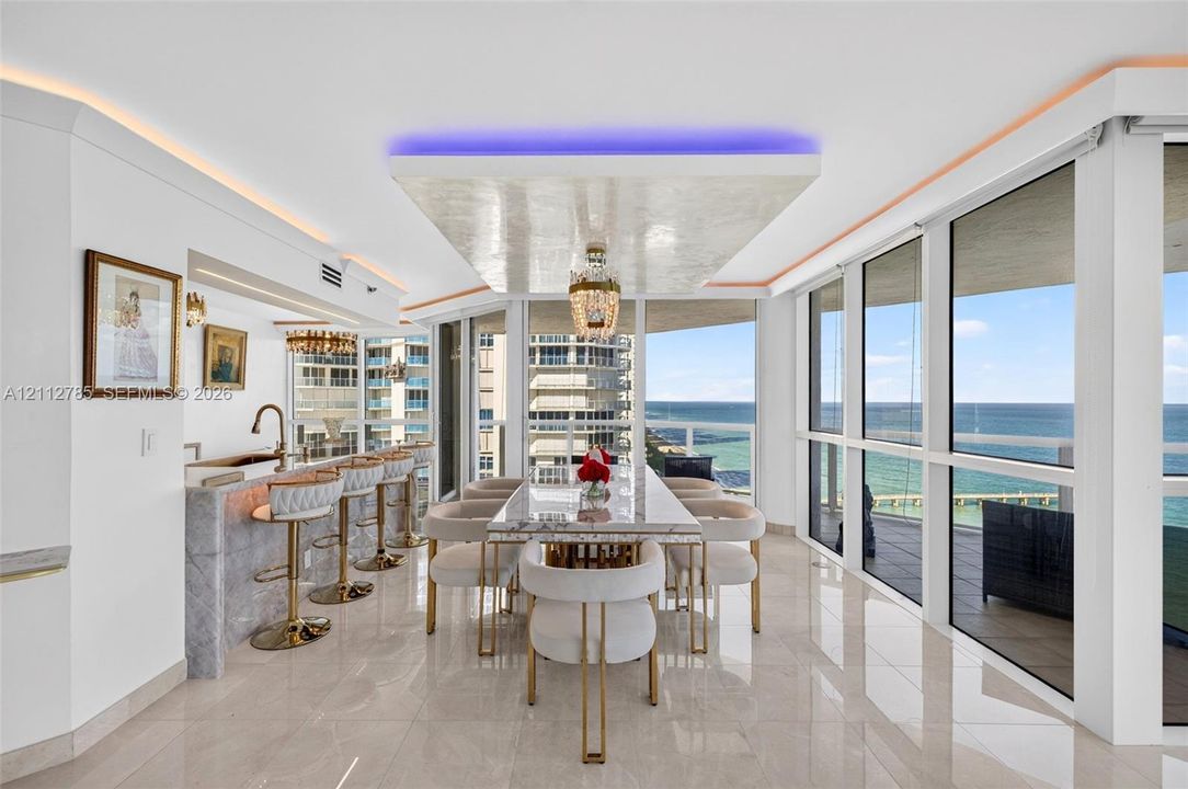 Floor-to-ceiling windows with ocean exposure, polished stone flooring, custom coffered ceiling with integrated accent lighting, wet bar with stone-surround, and a crystal chandelier