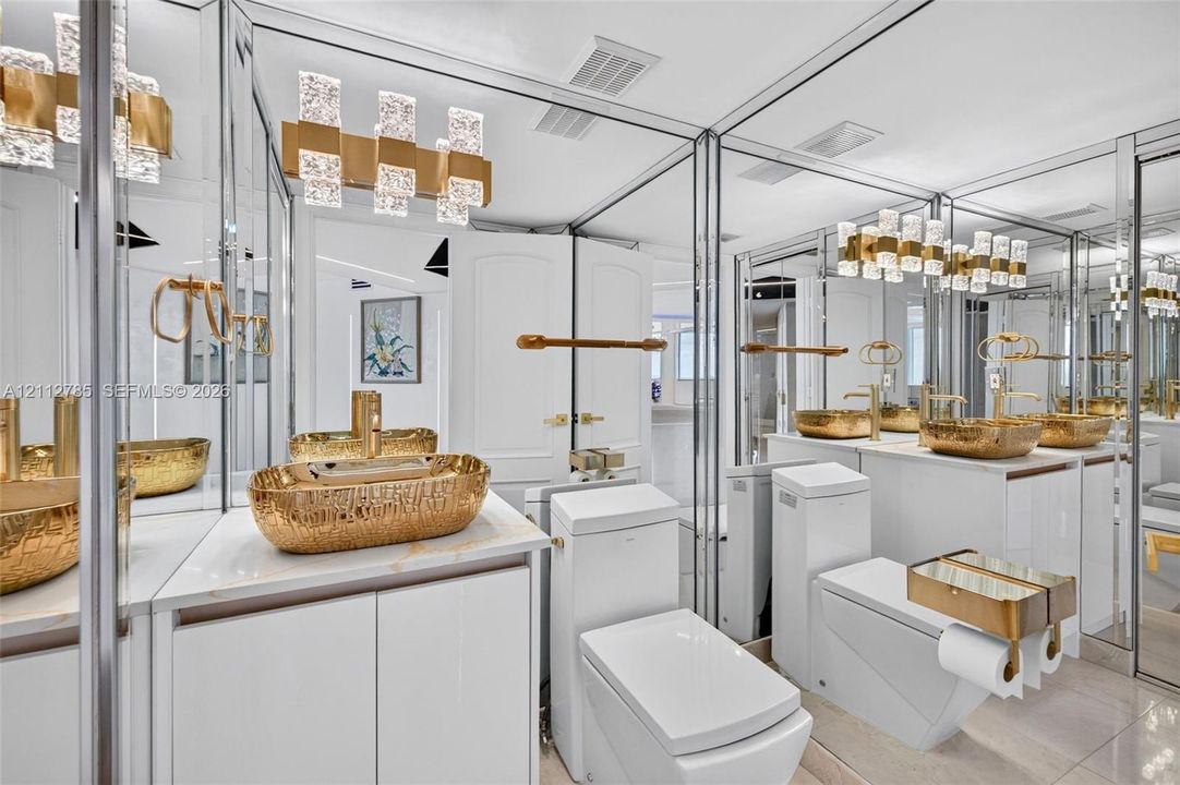 Bathroom featuring dual vanities with gold-tone hammered vessel sinks, quartz countertops, floor-to-ceiling mirrored walls, and tiered crystal-accented light fixtures