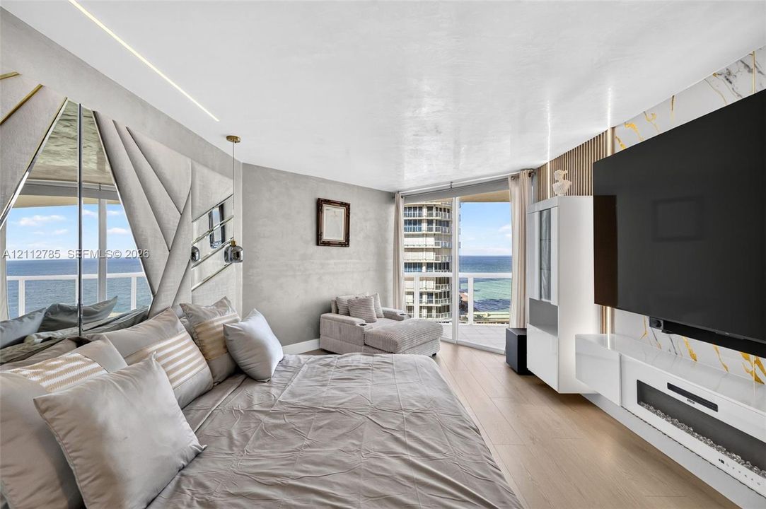 Wood-finish flooring, floor-to-ceiling windows with coastal scenery, accent wall with decorative mirror panels, wall-mounted linear fireplace, and integrated wood-slat wall paneling