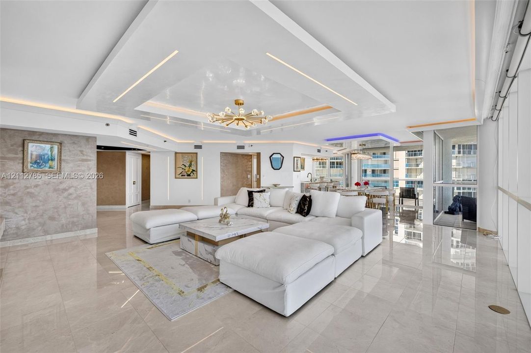 Open-concept living area featuring a custom recessed tray ceiling with integrated lighting, polished stone-finish flooring, floor-to-ceiling windows, and a textural wall finish