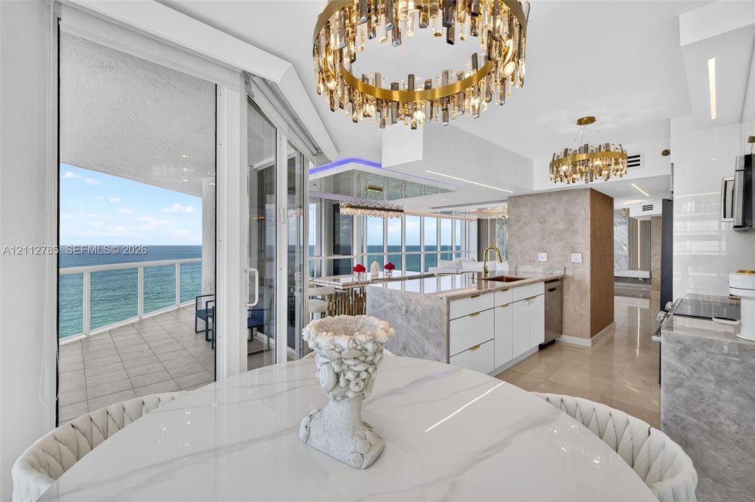 Open-concept kitchen featuring an island with stone-finish countertops, custom white cabinetry, gold-tone hardware, multi-tiered crystal statement chandeliers, and floor-to-ceiling glass doors opening to a tiled balcony