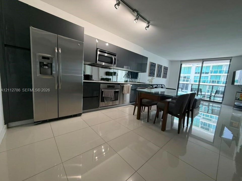 Kitchen featuring high-gloss dark cabinetry, stainless steel appliances, mirror backsplash, light-colored tile flooring, and track lighting