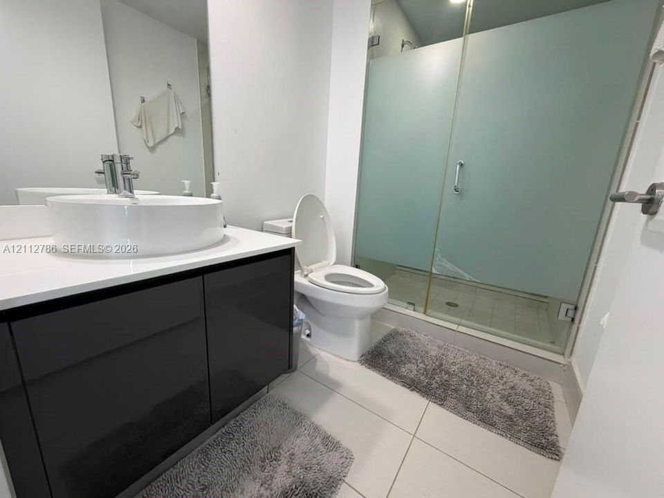 Bathroom featuring a high-gloss dark vanity, white composite countertop with a circular vessel sink, chrome faucet, and a walk-in shower with frosted glass enclosure