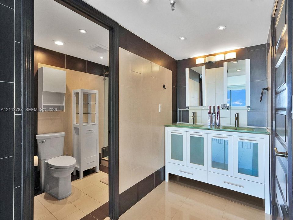 Bathroom featuring a dual-sink vanity with frosted glass cabinet doors, light-toned tile flooring, dark and cream-colored wall tile, and built-in storage cabinetry