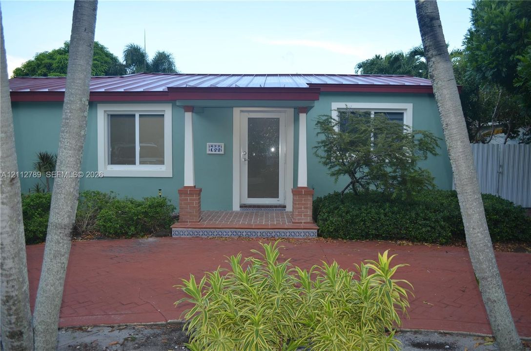Single-story exterior featuring a metal roof, covered entryway with brick-based columns, decorative tile riser, and brick-patterned hardscaping