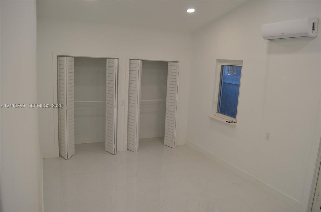 Interior room featuring light-toned flooring, two closets with white louvered bifold doors, a wall-mounted climate control unit, and recessed ceiling lighting