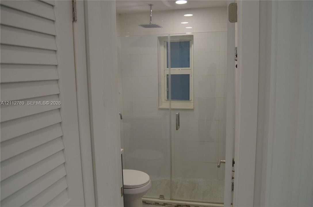 Full-height glass shower enclosure featuring a square rainfall showerhead, white tile wall surround, recessed lighting, and louvered door entry