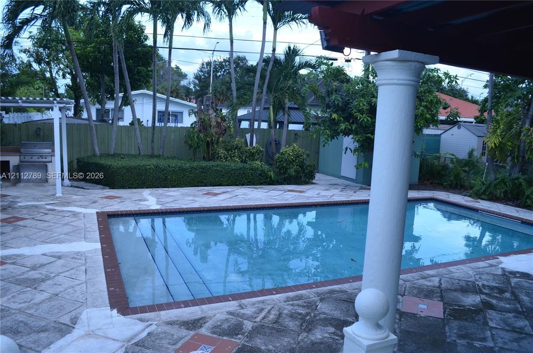 Rectangular swimming pool featuring brick coping and stone-finish decking, shaded pergola with a classical column, and built-in outdoor kitchen station