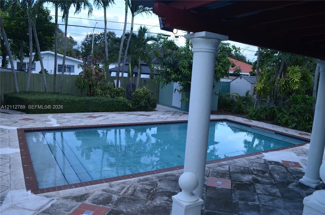 Rectangular swimming pool featuring a brick-accented border, stone patio, classic architectural columns, and mature palm trees
