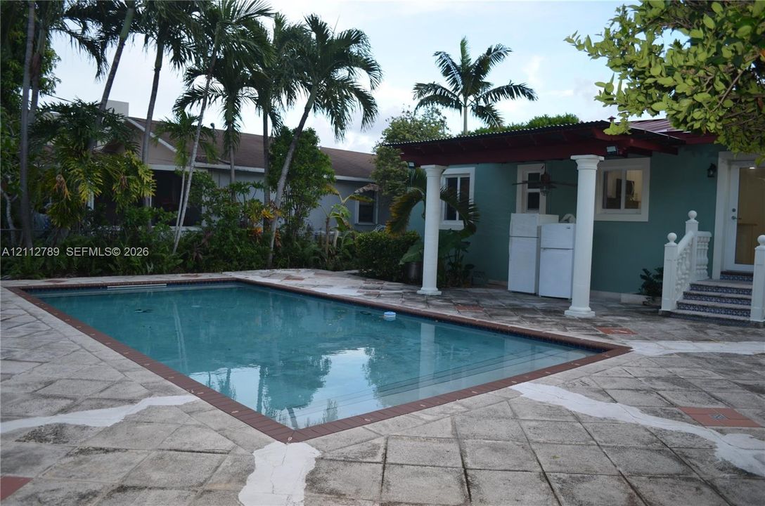In-ground swimming pool with masonry paver surround, covered patio with structural columns, and mature palm landscaping