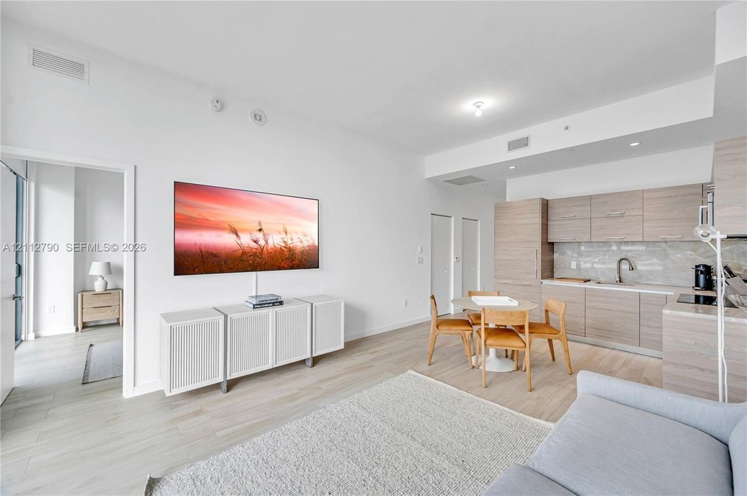 Open-concept living space featuring wood-finish flooring, light-toned flat-panel cabinetry, stone-finish backsplash, and recessed ceiling lighting
