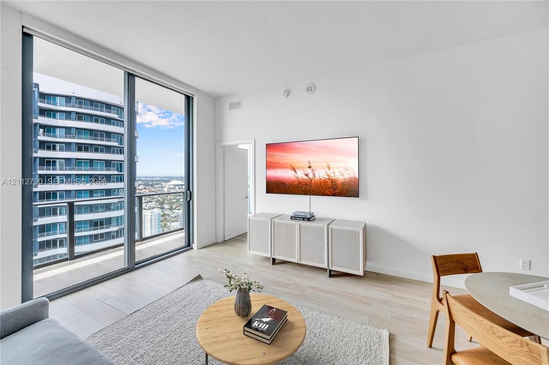 Floor-to-ceiling sliding glass doors, private balcony with metal railing, light-tone wood-finish flooring, and recessed ceiling lighting