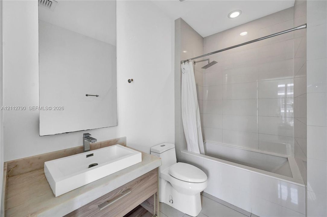 Bathroom featuring a wood-finish vanity with a rectangular vessel sink, solid-surface countertop, floor-to-ceiling white wall tile, and a built-in tub with a rainfall shower head