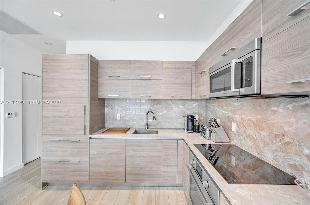 Modern kitchen featuring horizontal wood-finish cabinetry, stone-patterned backsplash, light-toned countertop, stainless steel microwave, and integrated refrigeration
