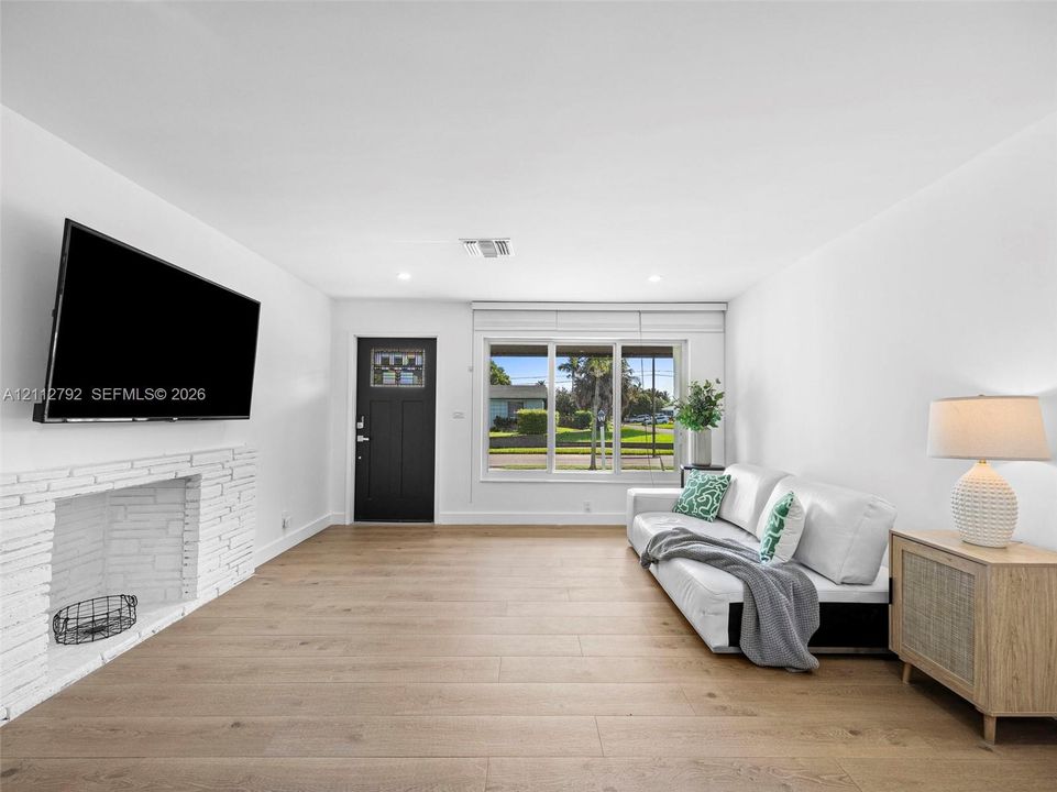 Living area featuring light wood-finish flooring, a white-painted brick fireplace, recessed lighting, and a multi-pane front door